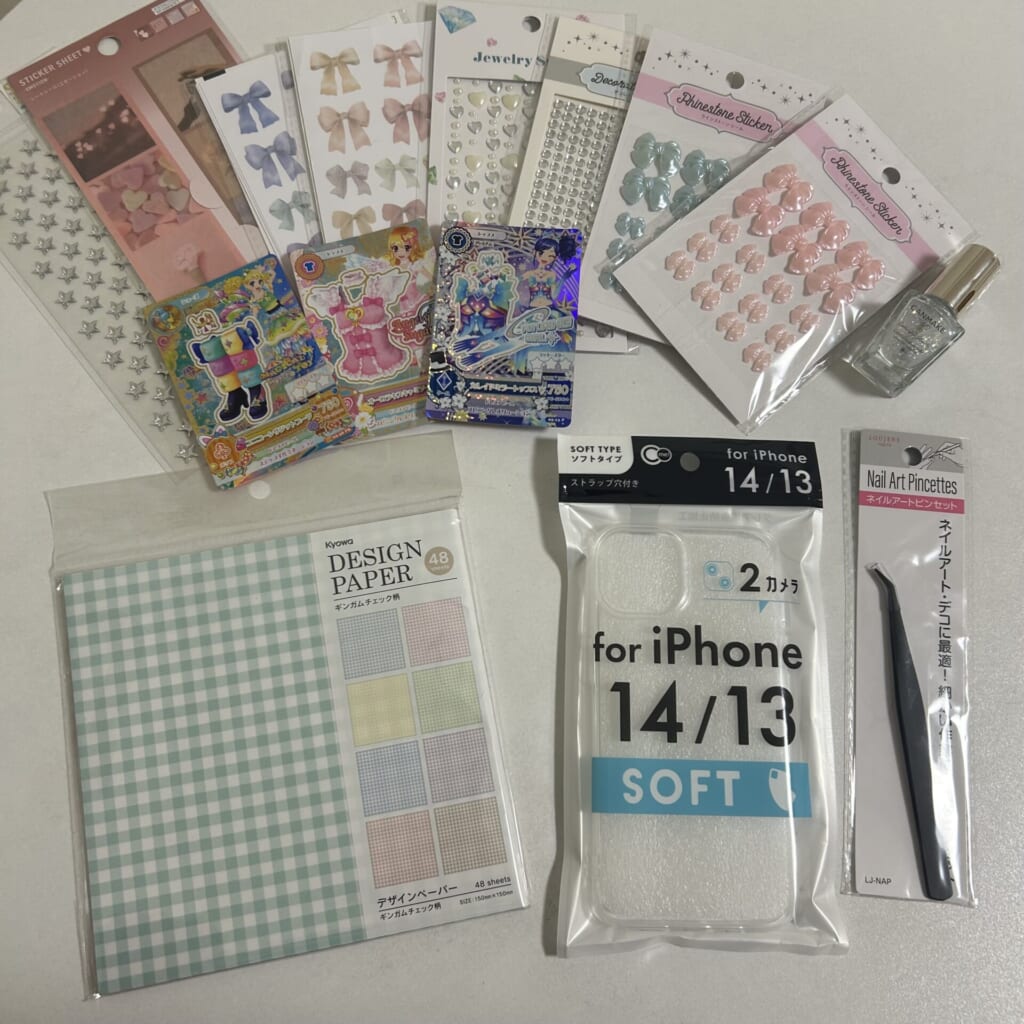 透明スマホケース・ピンセット・チェック柄の折り紙・リボンのシール・星のシール・ネイルのトップコート・アイカツカードなど、材料を並べて撮影した写真。