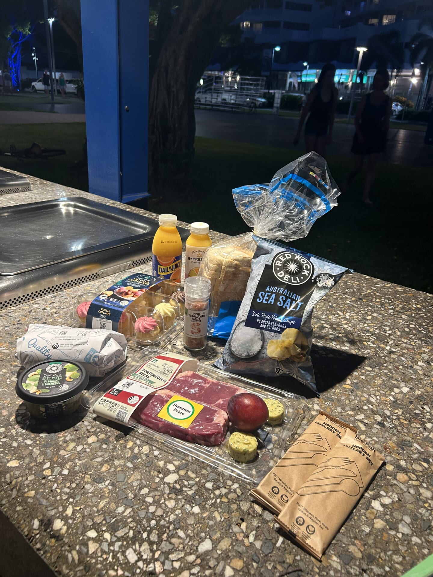 オレンジジュース、食パン、ポテトチップス、カップケーキ、塩、バジルソース、シーフードミックス、フルーツ、牛肉を並べた写真