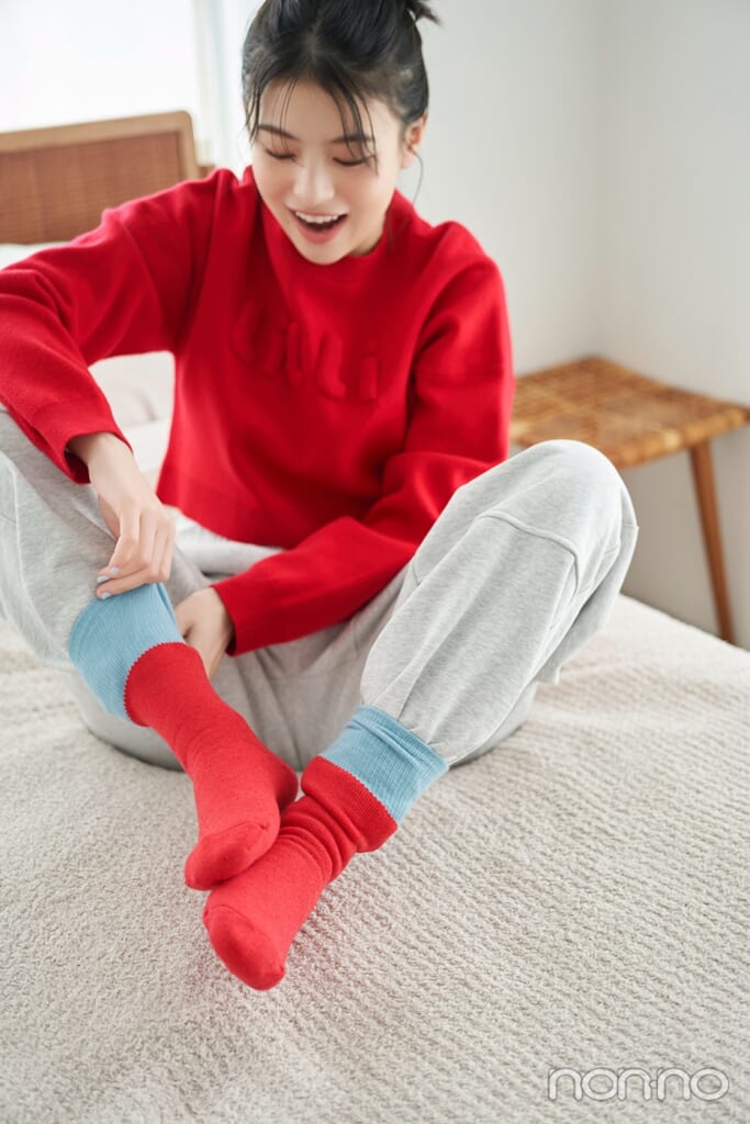 赤いニットにグレーのスエットパンツで靴下を履く出口夏希