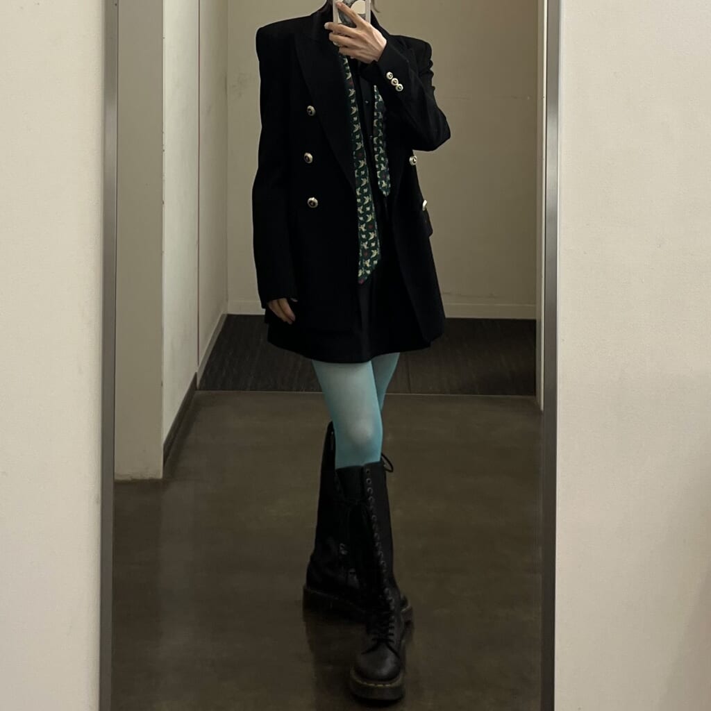 黒いジャケット 黒いスカート 黒いロングブーツに、ターコイズのカラータイツと緑のネクタイを合わせたモードコーディネート