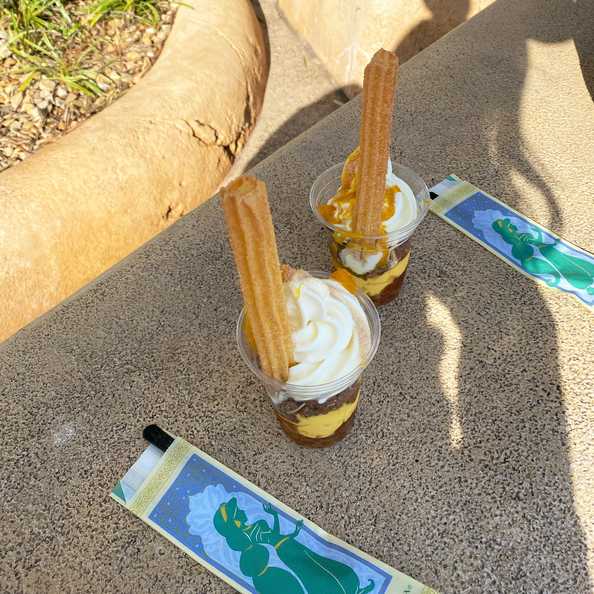 東京ディズニーシーで食べられるディズニーフード。季節限定メニューのサンデーパフェ。チャイゼリーとマンゴープリン、パイナップル、ソフトクリームが層になっていて、そこにチャイチュロスが刺さっています。アラビアンコーストにあるため、緑と水色、青を基調とした可愛いジャスミンのシルエットが描かれたスプーン入れが置いてあります。