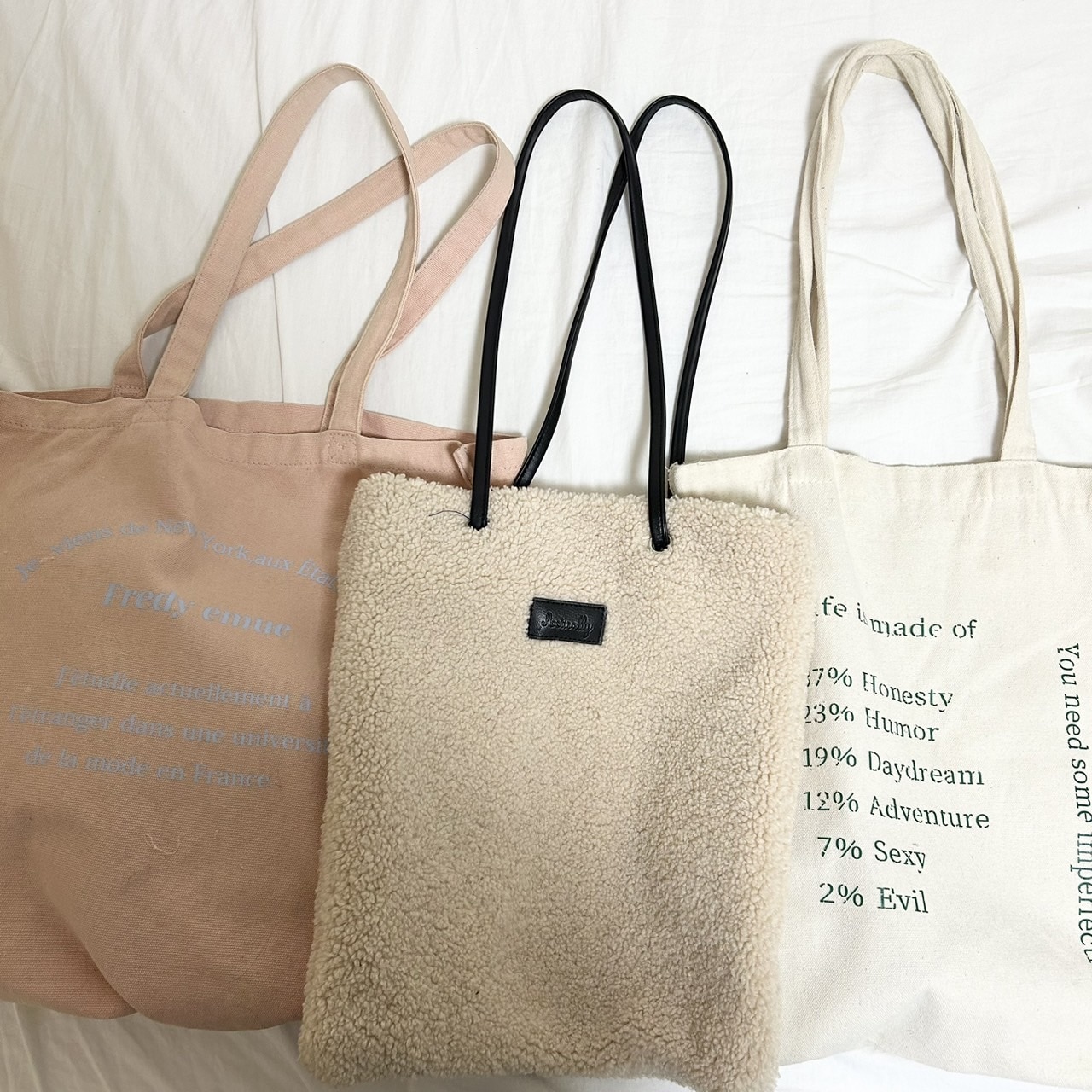三つのトートバッグの写真。白の生地に緑の英語が書かれたものと、薄ピンクの生地に水色の英語が書かれたもの、白のボア素材のものがある。