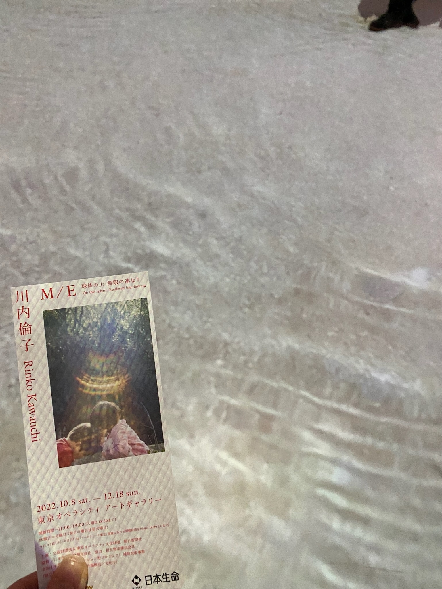 写真家・川内倫子さんの写真展「M/E 球体の上  無限の連なり」の様子。床には水面のようなプロジェクションマッピングが映し出され、左がわには写真展のチケットがあります。