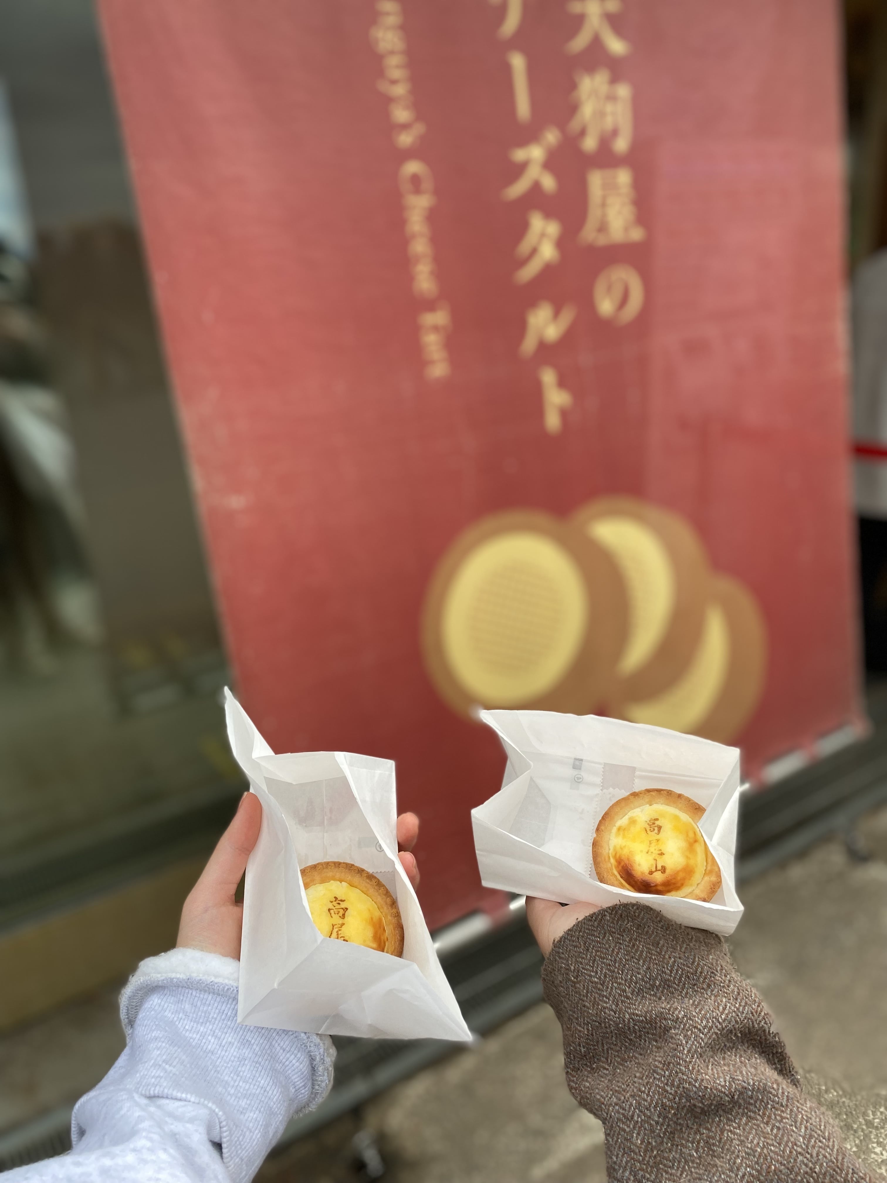 紅葉が綺麗な11月の高尾山に登山しに行った際に、ケーブルカー乗り場付近にあったチーズタルトのお店の前で撮ったチーズタルトの写真です。
奥には「天狗屋のチーズタルト」の文字と丸いチーズタルトのイラストが描かれた赤い布があり、手前には、手のひらサイズのチーズタルトが２つあります。